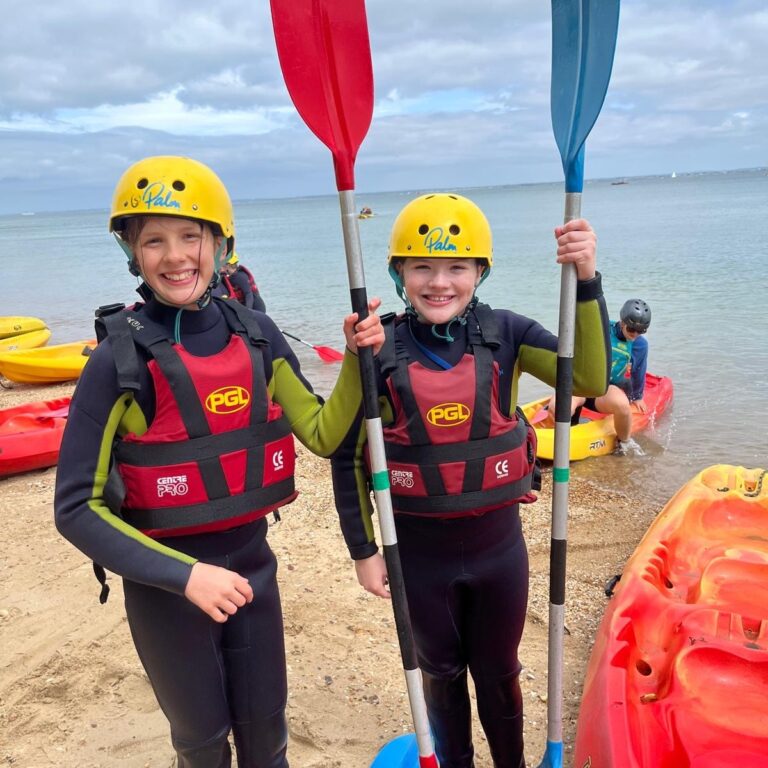Two children in wetsuits and yellow helmets stand on a sandy beach, holding red and blue paddles, wearing red life jackets with PGL logos, ready for kayaking.