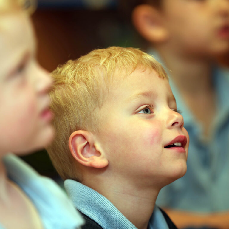Close-up of a blonde boy with blue eyes looking upward among a group of children in blue uniforms/shirts.