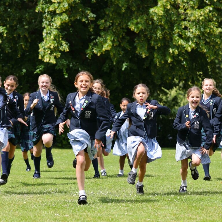 Group of schoolgirls in uniform running across a grassy field with trees in the background.
