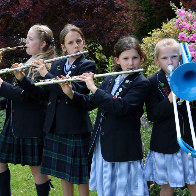 Four girls in school uniforms playing wind instruments outdoors in a garden with pink flowers behind them.