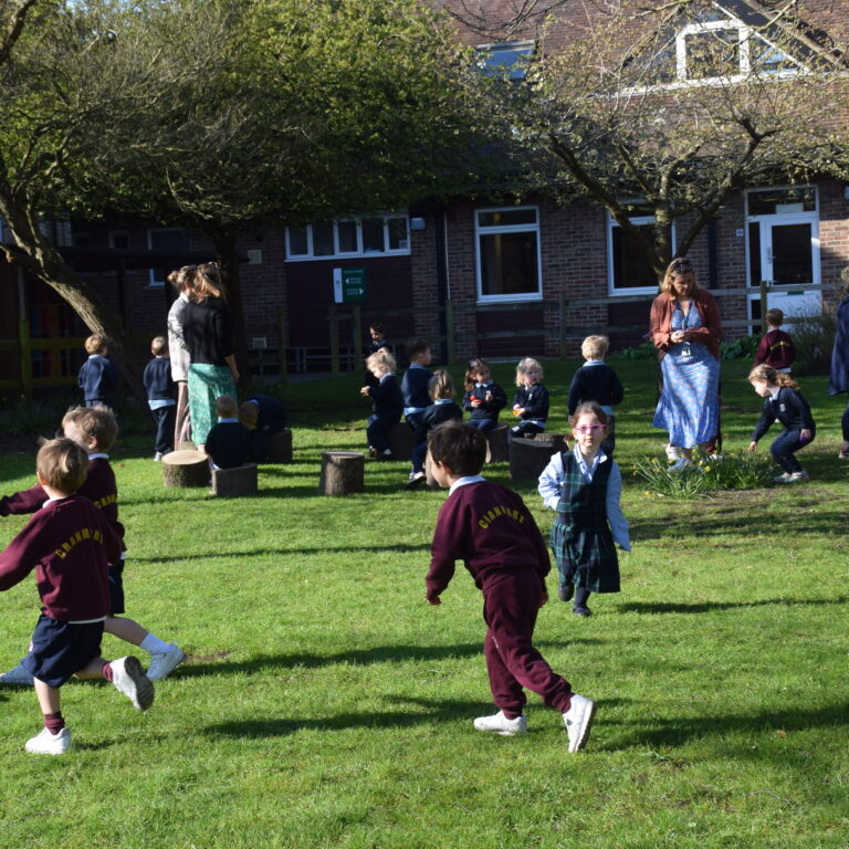 Group of children in maroon uniforms playing outside on a grassy schoolyard with adults supervising near a brick building.