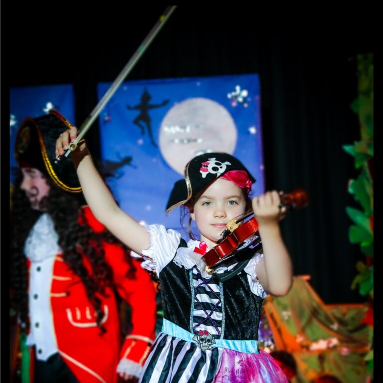 Young girl in a pirate costume plays a violin on a stage with a blue moon backdrop behind her.