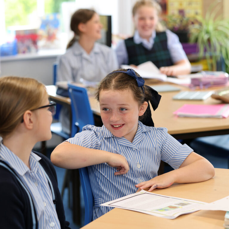 Student in a striped shirt with a blue hair bow sits at a desk, smiling, in a bright classroom with peers in the background.