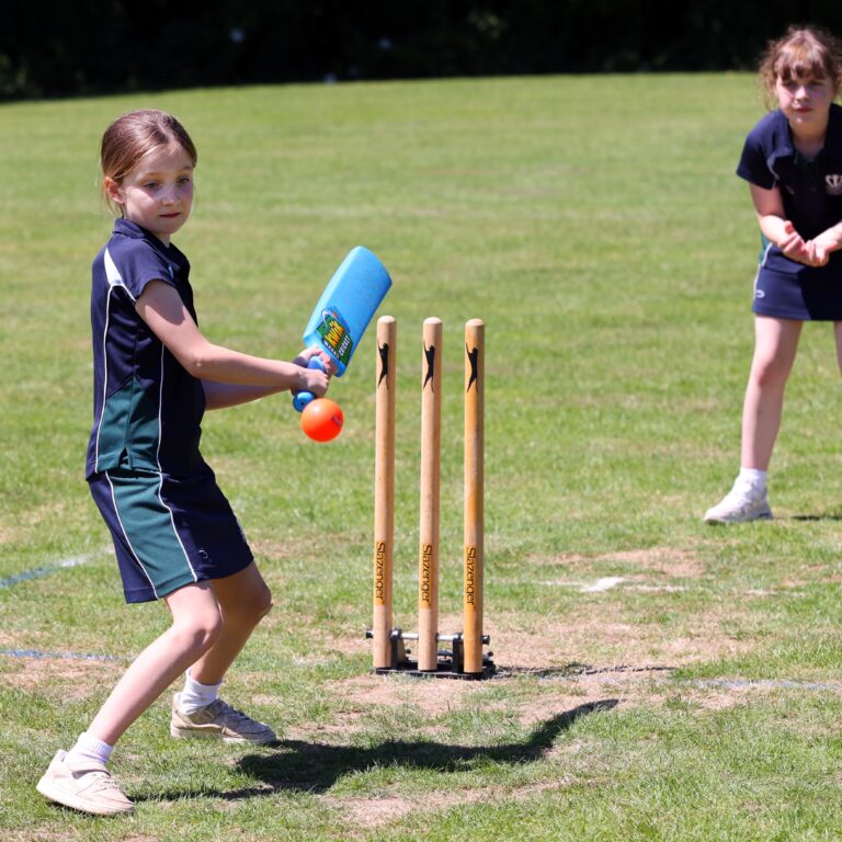 Young girl in a navy sports uniform swings a blue cricket bat at practice, with wooden stumps nearby on a sunny field.