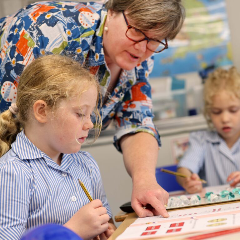 Teacher leaning in to help a young girl with a color-coded classroom activity at a desk in a bright classroom.