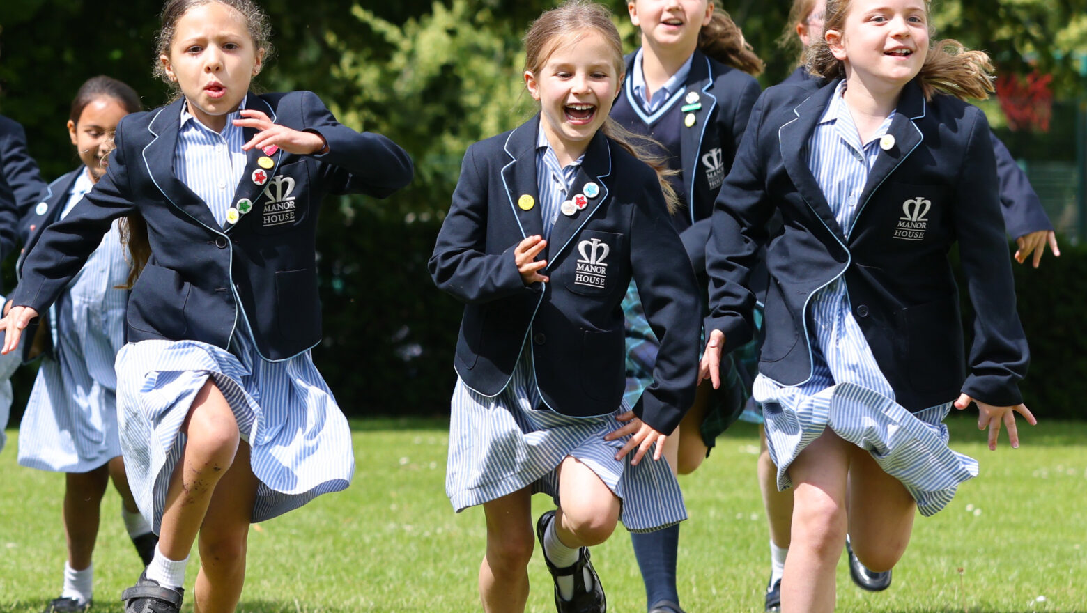 Group of girls in navy blazers and striped skirts running across a sunny grassy field with trees in the background.