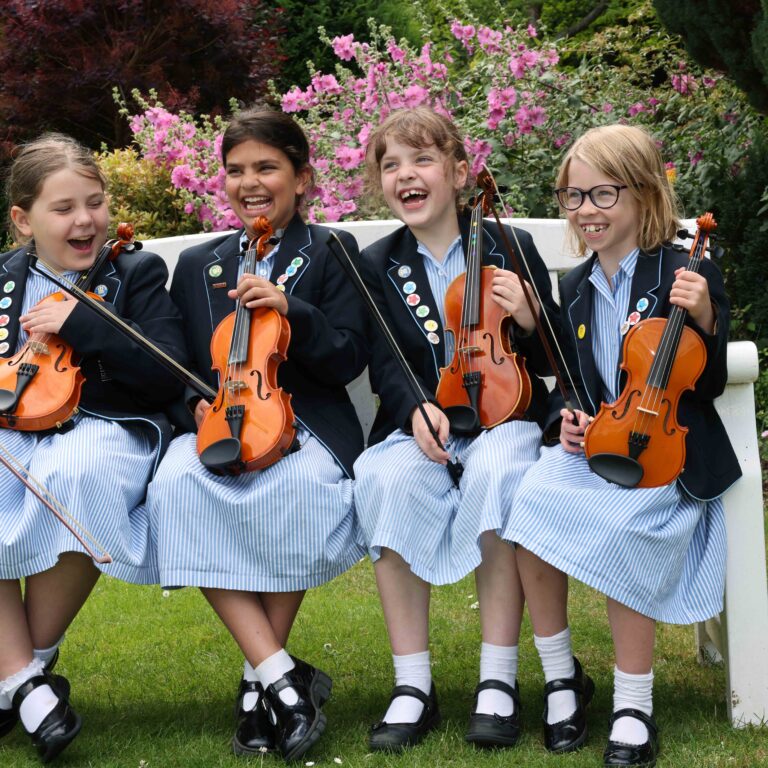 Four girls in school uniforms sitting on a bench with violins, laughing outdoors in a garden.