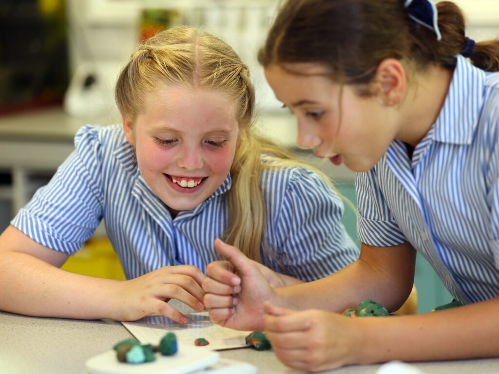 Two girls in blue-striped shirts work together shaping colorful clay on a classroom table.