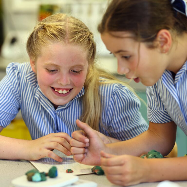 Two girls in blue-striped shirts work together shaping colorful clay on a classroom table.