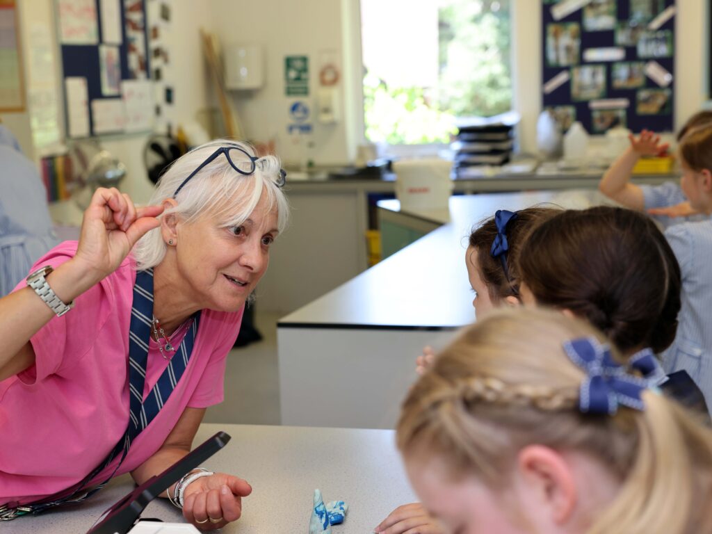 Older woman with glasses leaning toward a group of children at a classroom table, engaging in an activity.