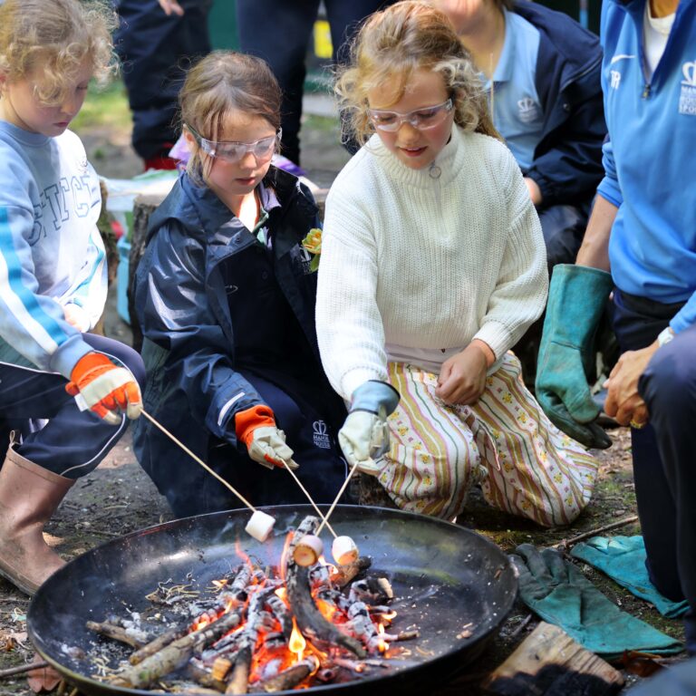 Children gathered around a small fire pit, roasting marshmallows on skewers with protective eyewear and gloves.
