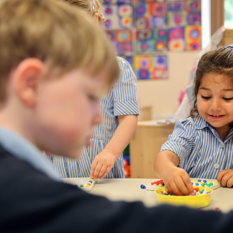 Two children in blue-striped uniforms sit at a table, arranging colorful beads on a bead board in a classroom backdrop of bright art displays.
