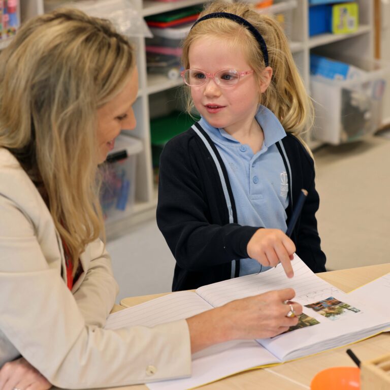 Adult and young student sit at a table, sharing a notebook as they write and discuss together in a classroom.