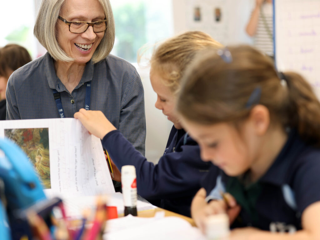 An older woman with glasses smiles as she sits beside a child, helping with a worksheet in a bright classroom.