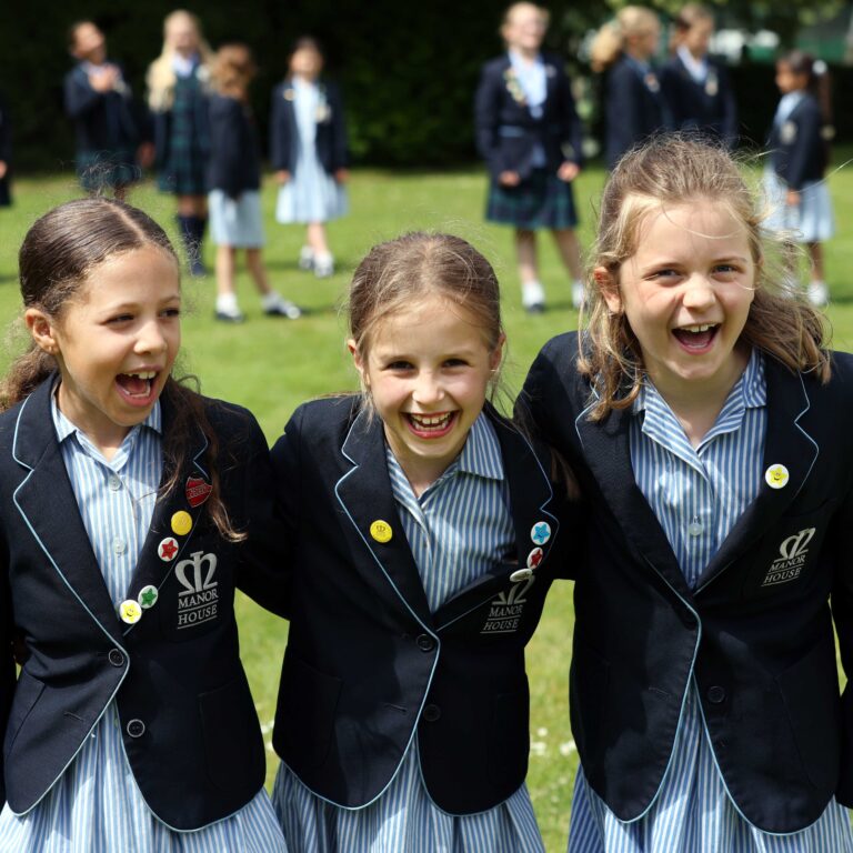 Three smiling schoolgirls in navy blazers with striped uniforms posing outdoors on a grassy field, with more students blurred in the background.