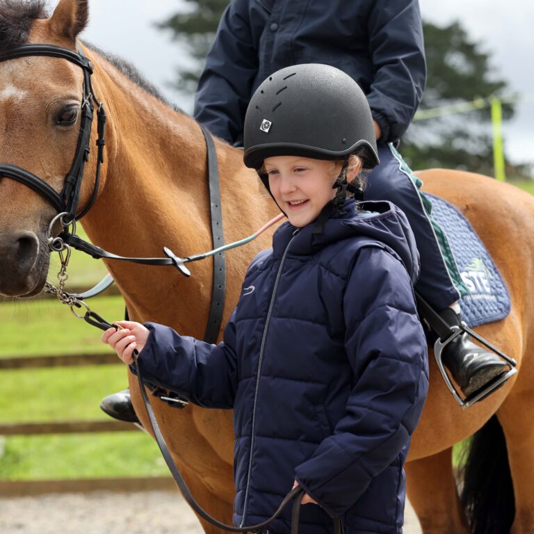 Child wearing a black riding helmet and navy jacket beside a brown horse with a bridle in a paddock.