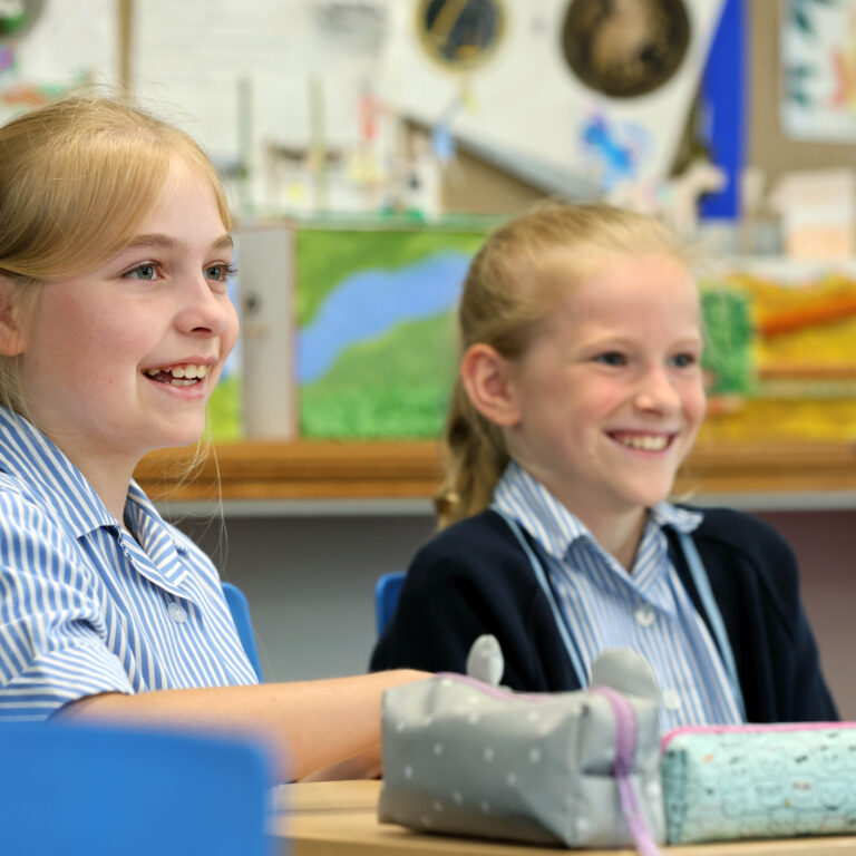Two smiling schoolgirls in blue-and-white striped uniforms sit at a classroom desk with colorful posters and supplies in the background.