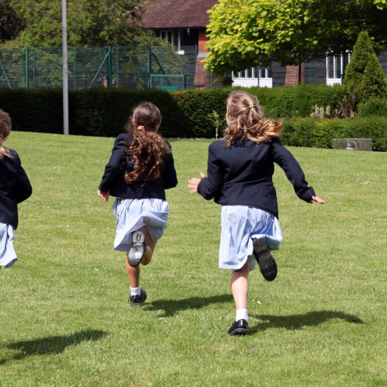 Three schoolgirls in navy blazers running across a grassy field, backs to the camera.
