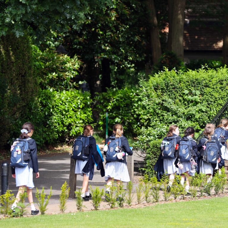 Group of schoolchildren in blue uniforms with backpacks walking along a path beside hedges and trees in a green garden setting.
