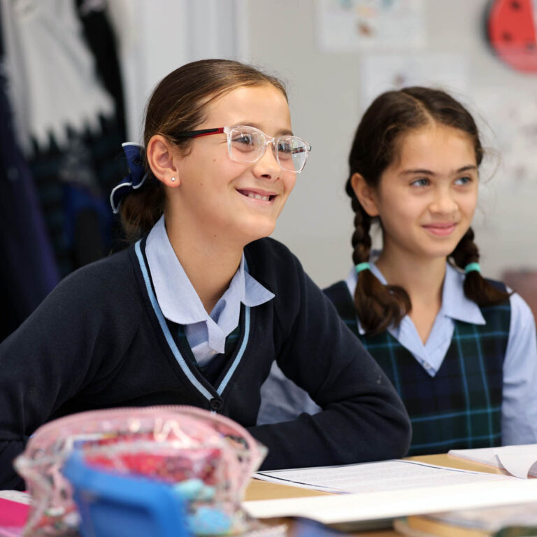 Two smiling schoolgirls in uniforms sit at a desk in a classroom.